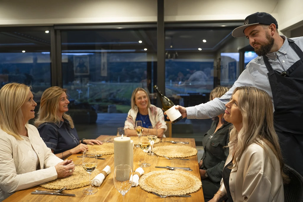 Chef Simon Lillico pouring wine for a group of female guests sitting at a dining table at a Kiwiesque Winter Food and Wine Experience